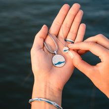 Laden Sie das Bild in den Galerie-Viewer, Zwei Hände mit Kette, Anhänger, Ring und Armband aus der Kollektion „Strandwellen“ – mit echtem Strandsand® und blauem Steinsand, aufgenommen vor dunklem Wasser