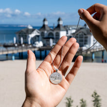 Laden Sie das Bild in den Galerie-Viewer, Hand hält runden Silberanhänger mit Motiv der Seebrücke Sellin vor realer Kulisse am Ostseestrand; Motiv aus Strandsand und Silberlinien eingearbeitet.
