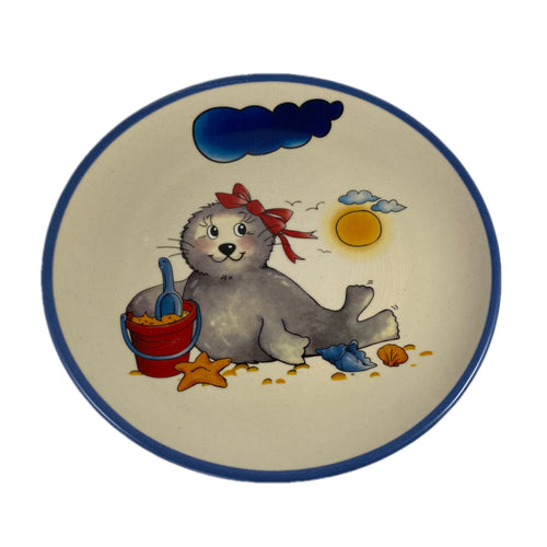Keramikteller mit Robbe. Die Robbe hat eine rote Schleife am Kopf und vor ihr befindet sich ein Buddeleimer 