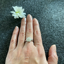 Laden Sie das Bild in den Galerie-Viewer, Ring Sandspirale von DUR Schmuck, getragen an einer Frauenhand auf grauem Untergrund und einer weißen Blüte