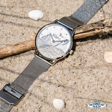 Laden Sie das Bild in den Galerie-Viewer, Edelstahl-Uhr „Strandwellen Rügen“ von DUR Schmuck, Zifferblatt mit echtem Strandsand, Wellenmotiv, Kreidefelsen, Segelschiff und Möwe, silbernes Meshband, fotografiert im Sand mit Muscheln.
