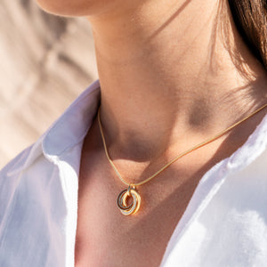 Anhänger „Marina“ aus vergoldetem 925er Silber mit drei ineinander verschlungenen Ringen, gefüllt mit Strandsand, Bernsteinsand und vergoldetem Silber, getragen an einer goldfarbenen Schlangenkette zu weißer Bluse.